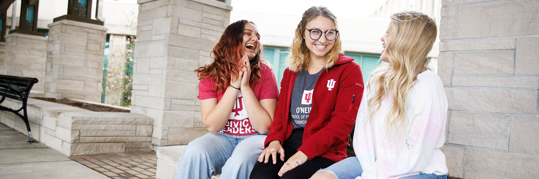 Three students laugh while seated on limestone bench. 